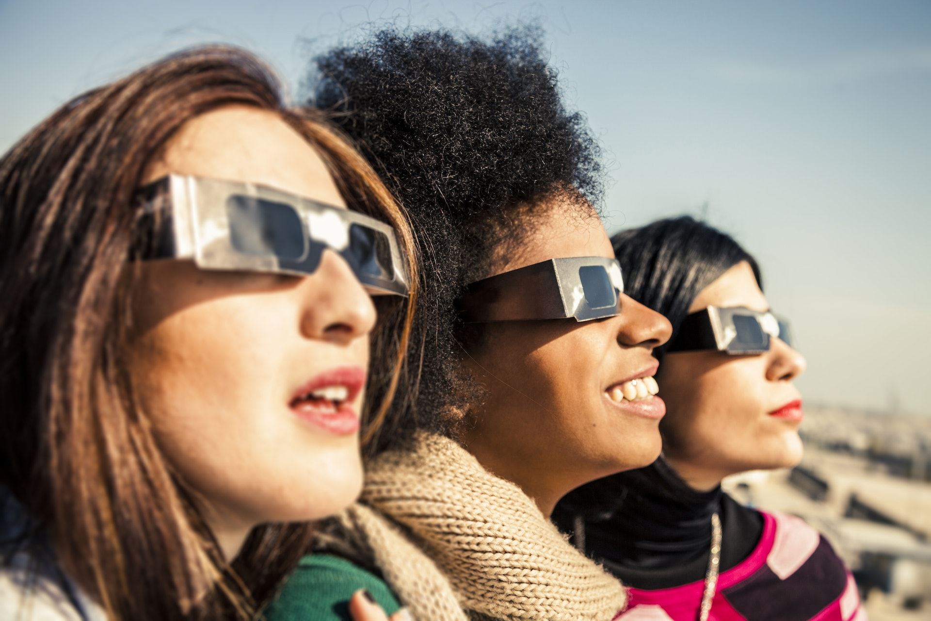 Three women wearing protective glasses look up at the sky during a solar eclipse