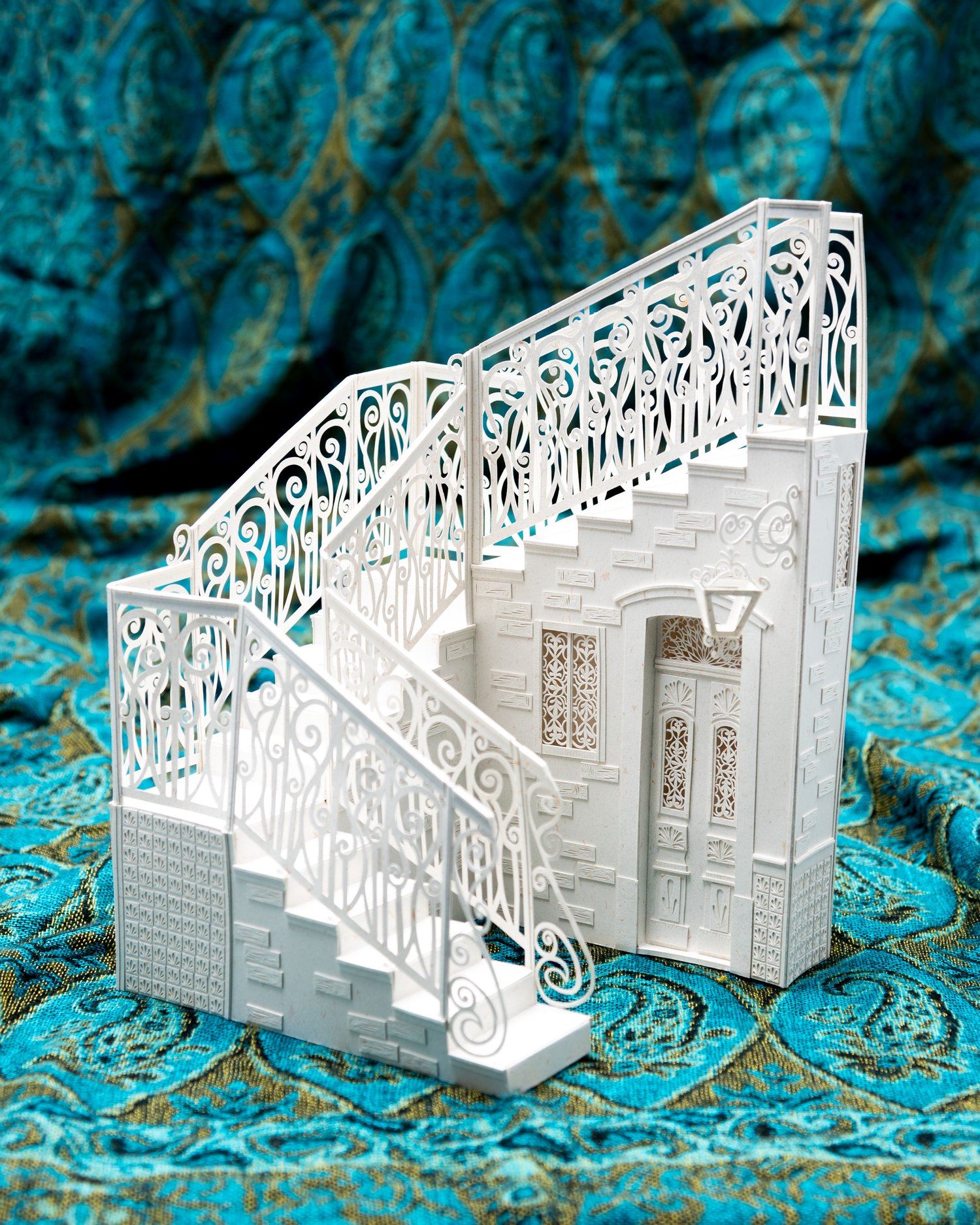 An architectural sculpture of a staircase made of paper, photographed on a teal paisley background.