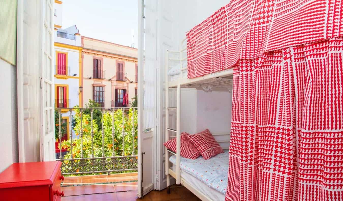 Bunk beds with red checkered privacy curtains, with an open door to a terrace showing the brightly colored buildings of Sevilla, Spain in the background